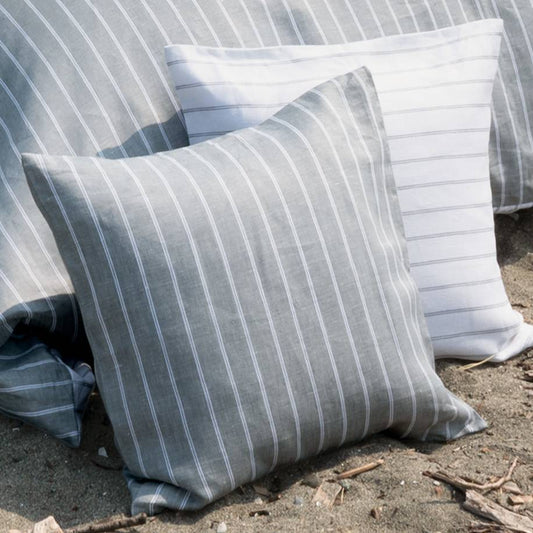 Two striped throw pillows in a linen-like fabric, one in gray and white and one in white and gray, resting on sandy ground outdoors.