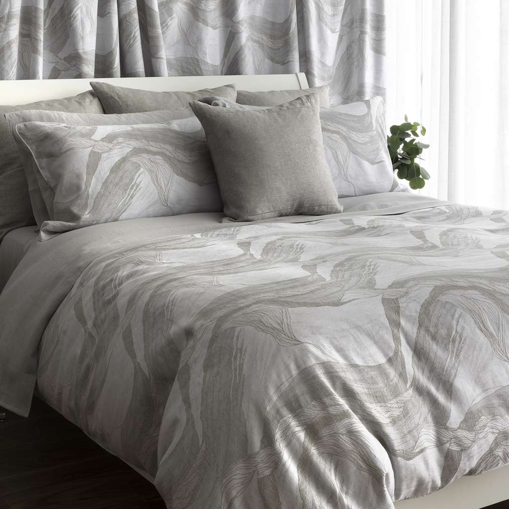 A neatly made bed with a light gray-and-white abstract wavy-patterned duvet cover and matching pillow shams, styled with a solid light gray accent pillow in a bright bedroom setting.