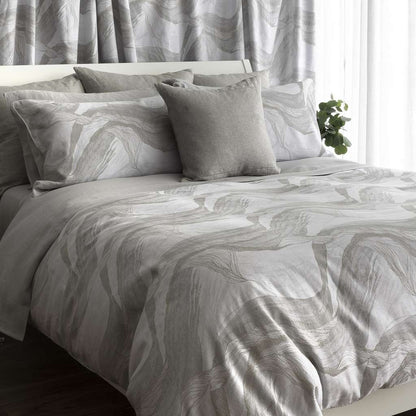 A neatly made bed with a light gray-and-white abstract wavy-patterned duvet cover and matching pillow shams, styled with a solid light gray accent pillow in a bright bedroom setting.