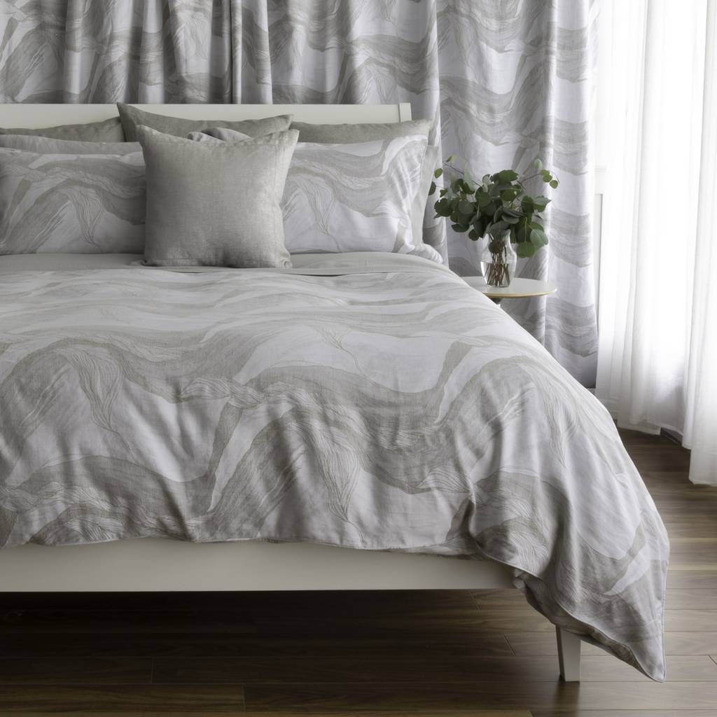 A neatly made bed with a light gray and white abstract wavy patterned duvet cover and matching pillow shams, styled in a bedroom setting with a small side table holding a green plant.