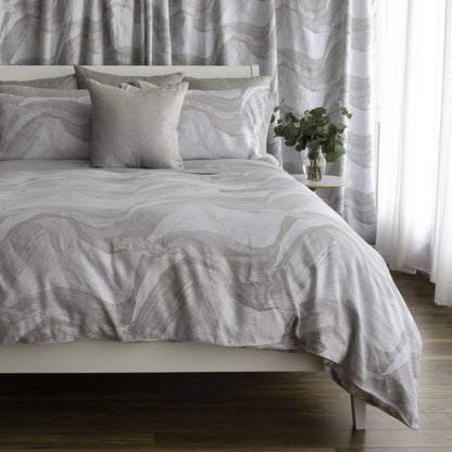 A neatly made bed with a light gray and white abstract wavy patterned duvet cover and matching pillow shams, styled in a bedroom setting with a small side table holding a green plant.