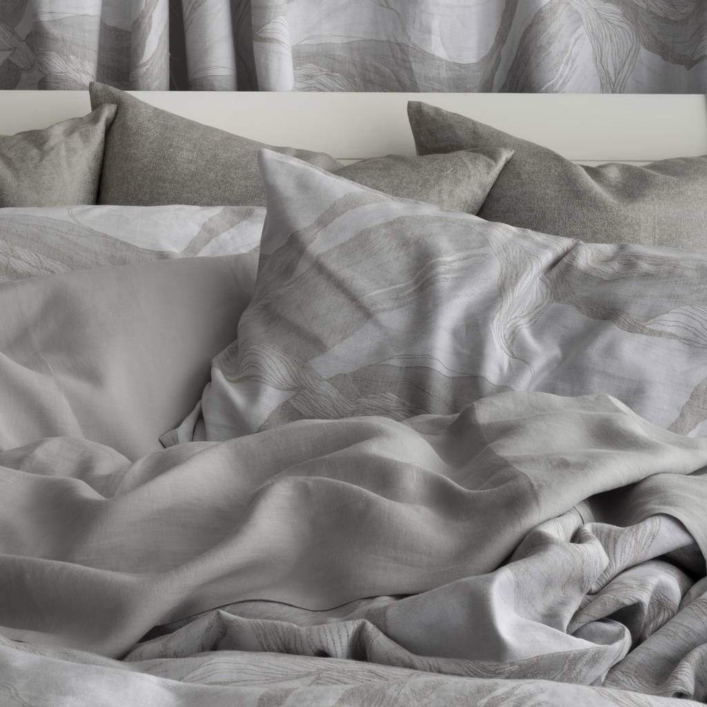A close-up of a bed made with light gray and white patterned bedding, including duvet cover, pillowcases, and matching pillows, with a white headboard and gray curtains in the background.