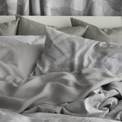 A close-up of a bed made with light gray and white patterned bedding, including duvet cover, pillowcases, and matching pillows, with a white headboard and gray curtains in the background.
