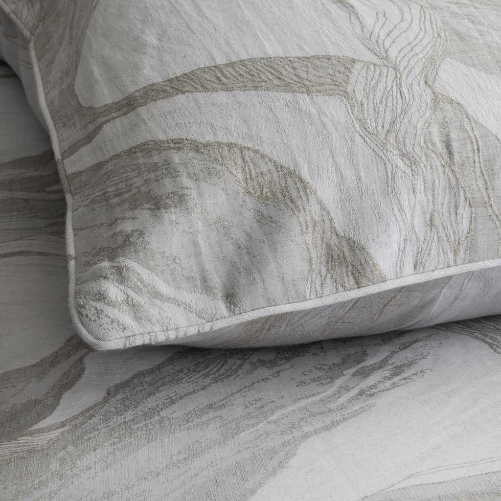 A close-up of a pillow sham with a textured, abstract botanical pattern in beige and gray on a white background, resting on matching patterned bedding.