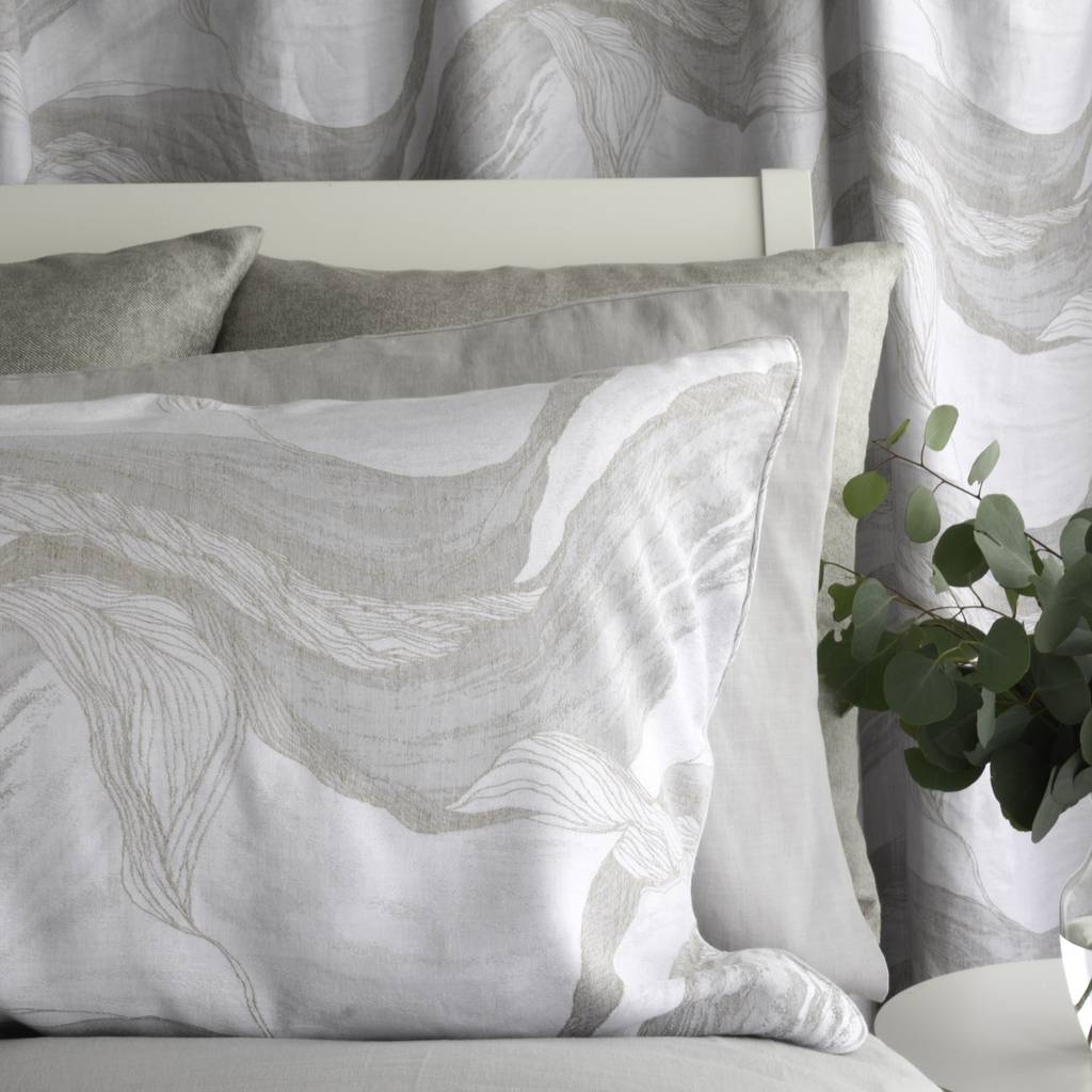 A styled bedroom scene showing a white pillow with a light gray abstract wave pattern, layered with solid gray pillows, in front of matching patterned curtains and a white headboard.