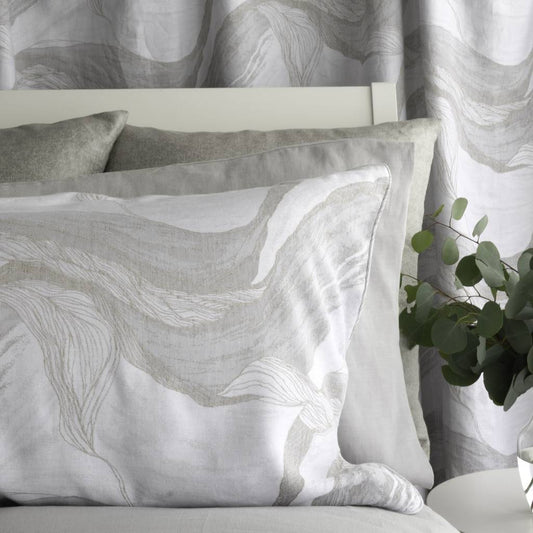 A styled bedroom scene showing a white pillow with a light gray abstract wave pattern, layered with solid gray pillows, in front of matching patterned curtains and a white headboard.