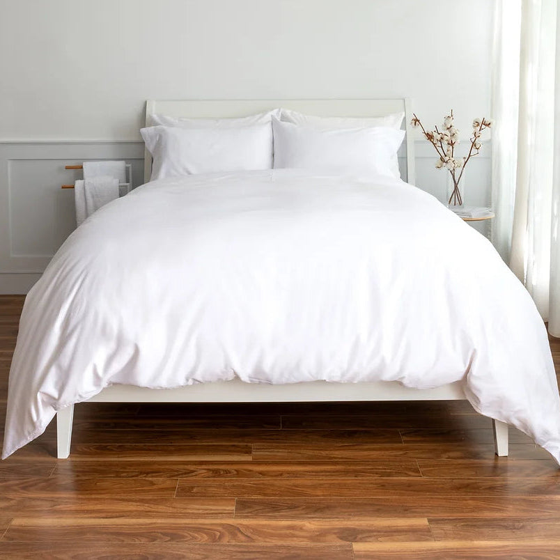 A neatly made bed with crisp white bedding, including a duvet cover and two pillows, set against a white headboard. The bed is positioned on a medium brown wooden floor in a bright bedroom with white walls and a small side table with a vase of branches.