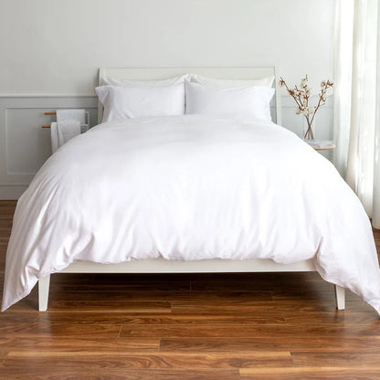A neatly made bed with crisp white bedding, including a duvet cover and two pillows, set against a white headboard. The bed is positioned on a medium brown wooden floor in a bright bedroom with white walls and a small side table with a vase of branches.