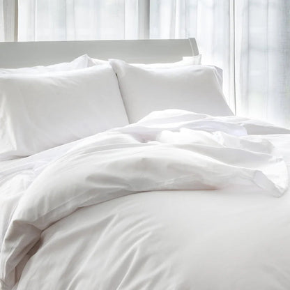 A white duvet cover with a silky appearance, displayed on a bed with pillows, in a room with a window behind.